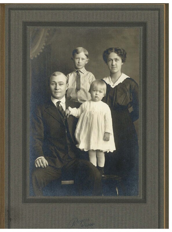 a black and white photo of a family of four: two adults and two children. The father is dressed in a three piece suit, and is seated on the left, and the mother is wearing a dress with a lace collar, and is standing on the right. The children are posed between them: a young boy standing and a toddler standing on the chair.