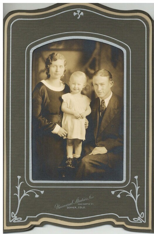 a sepia tone family portrait of a family of three: a mother stands on the left and a father sits on the right. Between them a little girl in a dress stands and smiles.