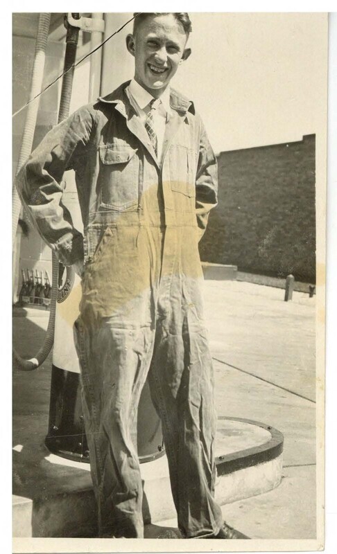 a black and white photo of a young man with a big smile, dressed in coveralls, hands on hips, seemingly standing in front of a gas pump.