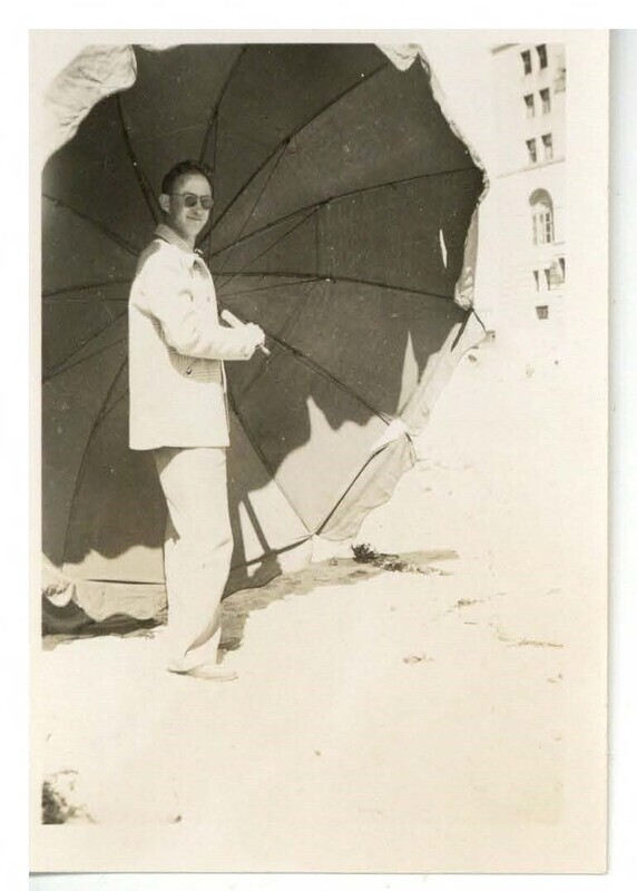 a black and white photo of a man in light casual clothing wearing sunglasses holding an oversized umbrella and grinning.