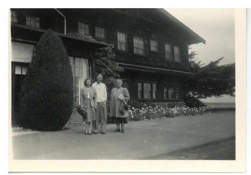 a black and white photo of a man and two women at a distance, standing in front of a large house.