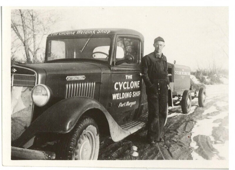 a black and white photo of a man in dark coveralls and a hat leaning against a large truck that reads 'The Cyclone Welding Shop Fort Morgan'. There is some snow on the ground.