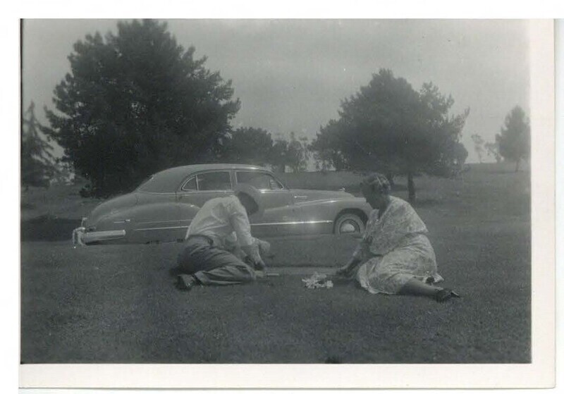 a black and white photo of a man and a woman sitting on the grass on a sunny day near a gravesite. The woman has flowers; the man istouching the gravestone. Behind them is a car and some trees.