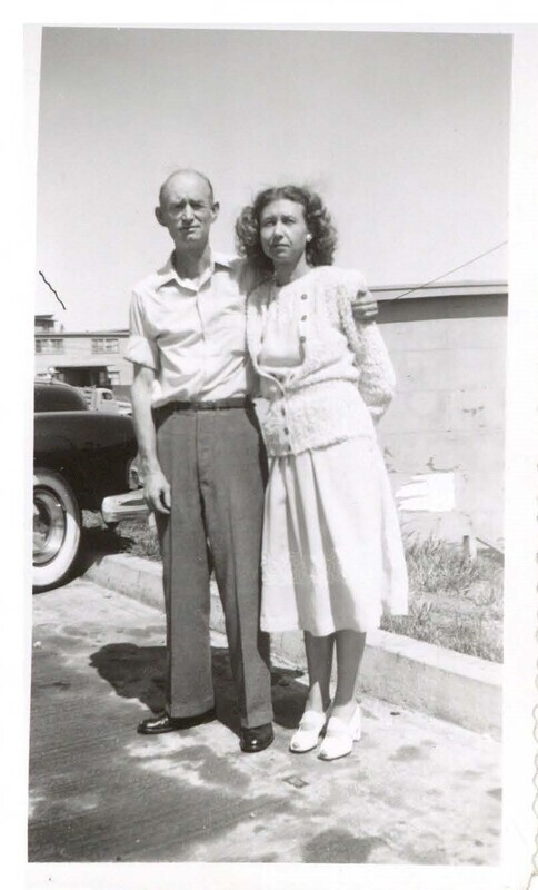 a black and white photo of two people standing near a curb: a balding man in a short-sleeve button up shirt with his arm around a woman with curly hair in a light dress and cardigan. They are squinting into the sun.