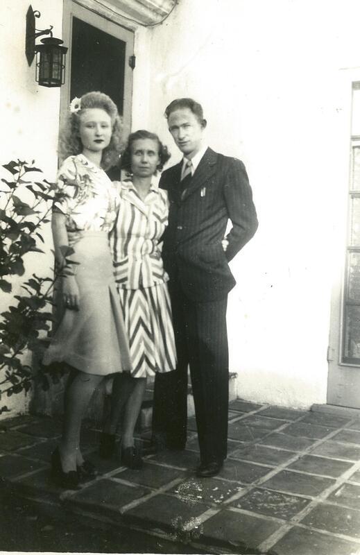 a black and white photo of a family standing outside their home. the teenage daughter is on the left. she is tall and thin with a flower in her blond hair. the mother stands in the middle with a striped dress and the father stands on the right in a pinstripe suit.
