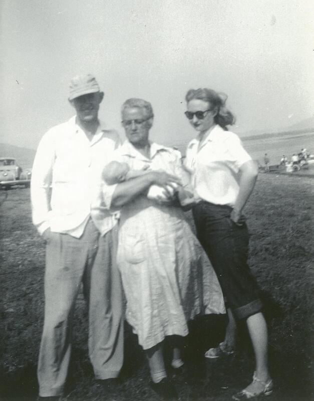 a black and white photo of a group of people standing outside on a windy day. the man is smiling, dressed in light clothes and a cap. the older woman is holding an infant, and the baby's mother is wearing sunglasses and stands with her hands on her hips.