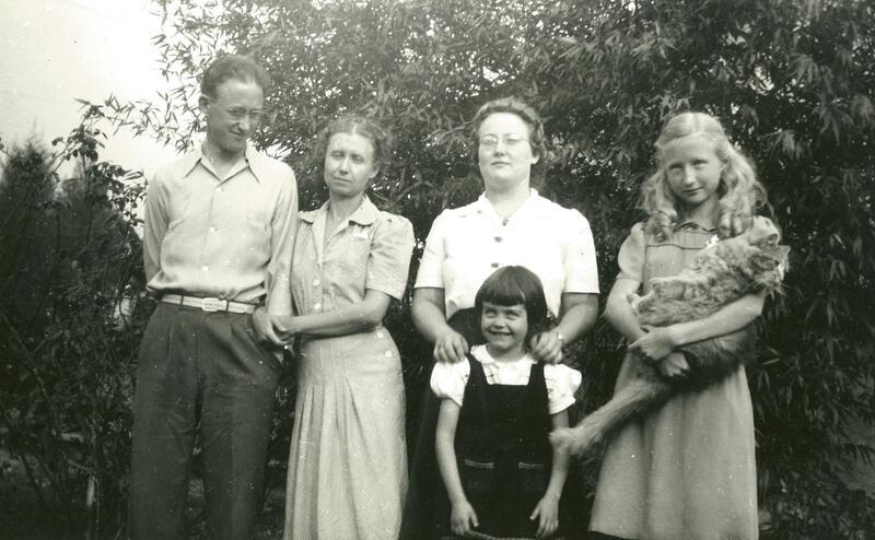 a black and white photo of a group of people standing outside, including: a man smiling at everyone, linking arms with a woman. Another woman stands behind a young girl, and a young teenager holds a longhair cat like a baby.