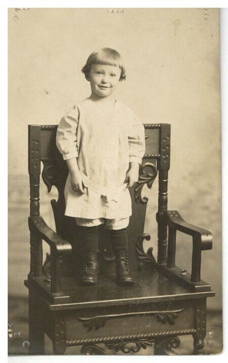 a black and white photo of a young boy smiling in a light-colored smock and black boots standing on a large ornate wooden chair.