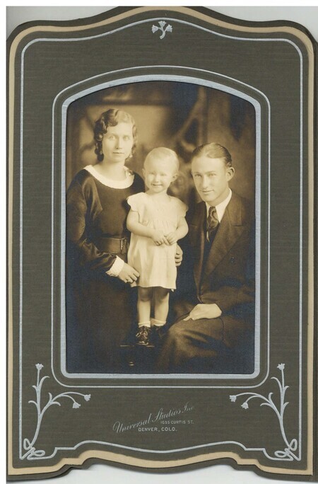 a sepia tone family portrait of a family of three: a mother stands on the left and a father sits on the right. Between them a little girl in a dress stands and smiles.