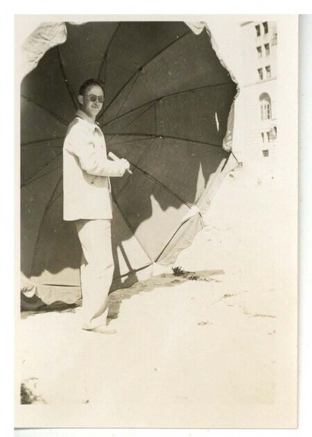 a black and white photo of a man in light casual clothing wearing sunglasses holding an oversized umbrella and grinning.