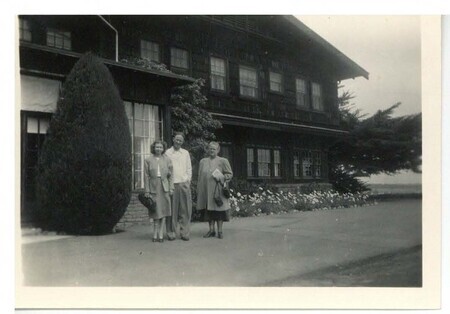 a black and white photo of a man and two women at a distance, standing in front of a large house.