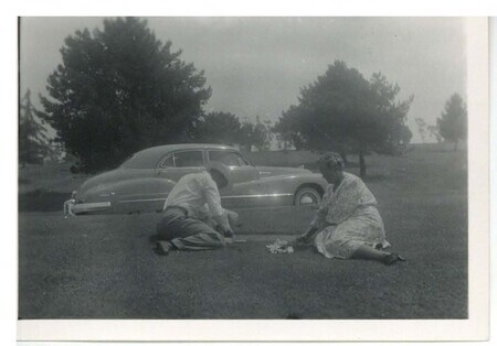 a black and white photo of a man and a woman sitting on the grass on a sunny day near a gravesite. The woman has flowers; the man istouching the gravestone. Behind them is a car and some trees.