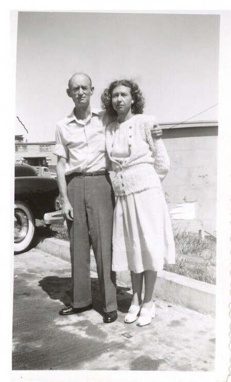 a black and white photo of two people standing near a curb: a balding man in a short-sleeve button up shirt with his arm around a woman with curly hair in a light dress and cardigan. They are squinting into the sun.