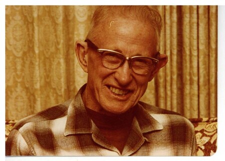 a color photo of an older man smiling with wayfarer glasses and a plaid shirt in front of gold curtains.