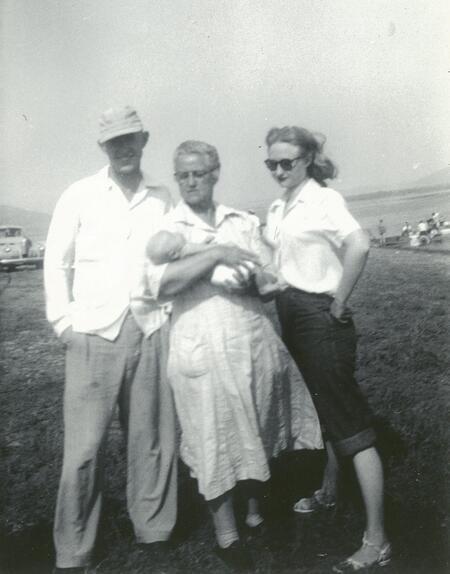 a black and white photo of a group of people standing outside on a windy day. the man is smiling, dressed in light clothes and a cap. the older woman is holding an infant, and the baby's mother is wearing sunglasses and stands with her hands on her hips.