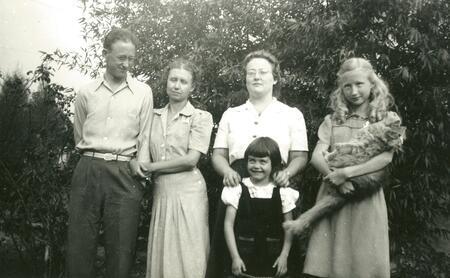 a black and white photo of a group of people standing outside, including: a man smiling at everyone, linking arms with a woman. Another woman stands behind a young girl, and a young teenager holds a longhair cat like a baby.